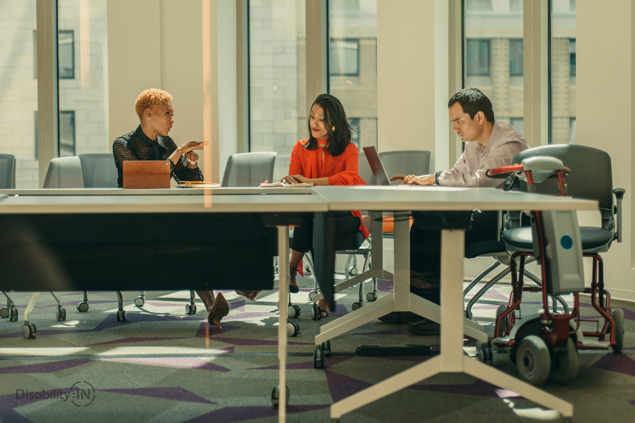 Three people sit at a conference table using laptops and sign language. A wheelchair is next to one person.